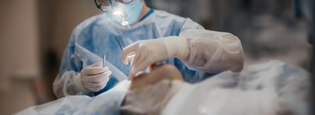 Masked medical professional wearing gloves and magnifying eyewear performs a procedure on a reclining patient.